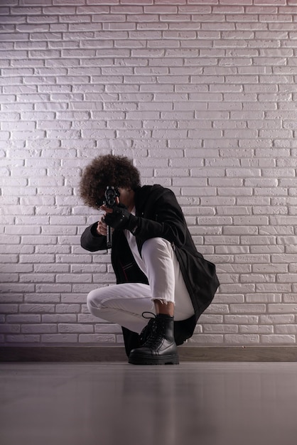 Un árabe con un peinado africano en elegante ropa de moda con una pistola de airsoft posa contra una pared de ladrillos