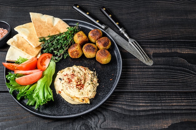 Arabische Küche Hummus Kichererbsen, Falafel, Fladenbrot und frisches Gemüse. Schwarzer hölzerner Hintergrund. Ansicht von oben. Platz kopieren.