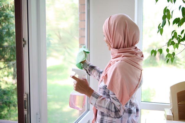 Arabische Muslimin mit bedecktem Kopf in einem Hijab wäscht Fenster, entfernt Flecken, sprüht Waschmittel und wischt mit einem Lappen ab und genießt einen allgemeinen Frühjahrsputz des Hauses