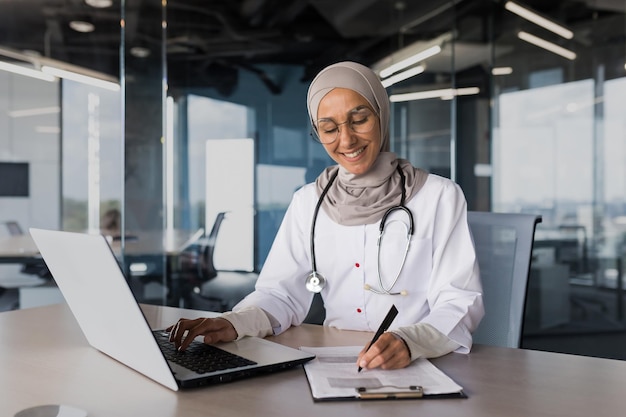 Arabische muslimische Frau in Hijab-Ärztin, die in einem modernen Klinikbüro mit Laptop-Arzt an Papierkram arbeitet