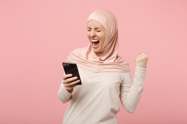 Arabische muslimische Frau in Hijab-leichter Kleidung, die isoliert auf rosafarbenem Hintergrund posiert. Menschen religiösen Islam Lifestyle-Konzept. Mock-up-Kopienbereich. Mit dem Handy, SMS-Nachricht eingeben, Siegergeste machen.