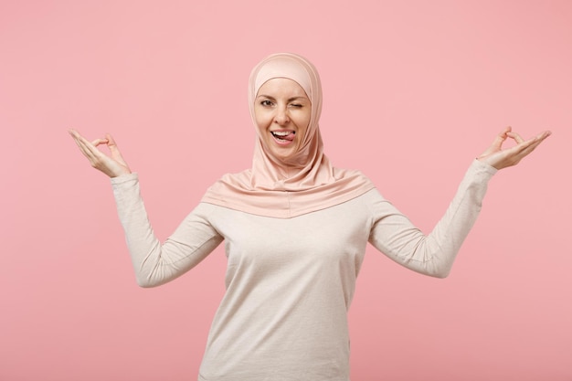 Arabische muslimische Frau in Hijab-leichter Kleidung posiert isoliert auf rosa Hintergrund. Menschen religiösen Islam Lifestyle-Konzept. Mock-up-Kopienbereich. Hände in Yoga-Geste halten, meditieren, Zunge zeigen.