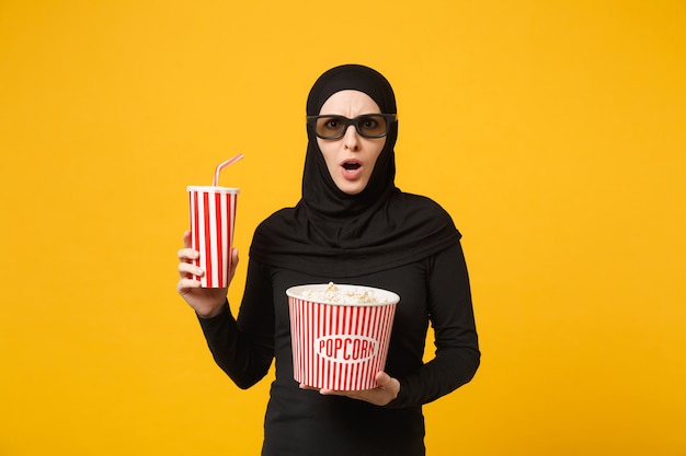 Arabische muslimische frau in hijab schwarzer kleidung 3d imax brille sehen film film halten popcorn, tasse soda isoliert auf gelbem wandporträt. Menschen Lifestyle-Konzept. .
