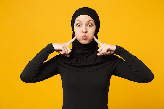 Arabische muslimische Frau in Hijab schwarzer Kleidung, die Zeigefinger auf blasende Wangen zeigt, isoliert auf gelber Wand, Porträt. Menschen religiösen Islam Lifestyle-Konzept.