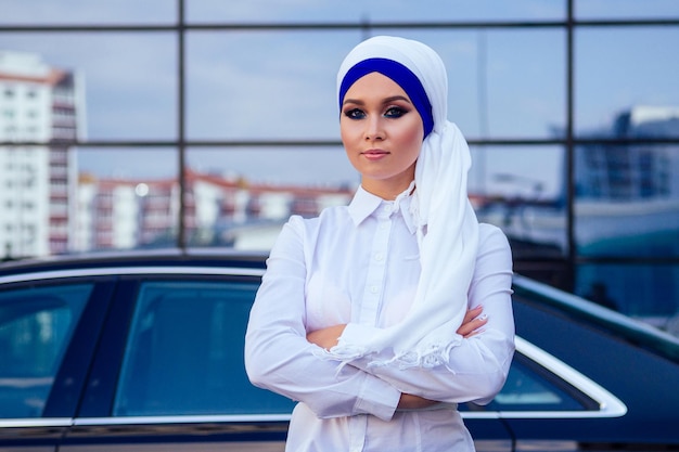 Arabische muslimische Geschäftsfrau Hijab mit Make-up, die vor ihrem Luxusauto auf der Straße vor einem Hintergrund von Wolkenkratzern steht Die Frau ist in eine stilvolle Abaya gekleidet