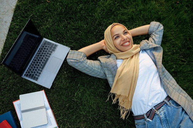 Arabische Studentin im Hijab, der auf dem Gras im Sommerpark, Draufsicht liegt. Muslimische Frau mit Büchern, die auf dem Rasen ruhen.