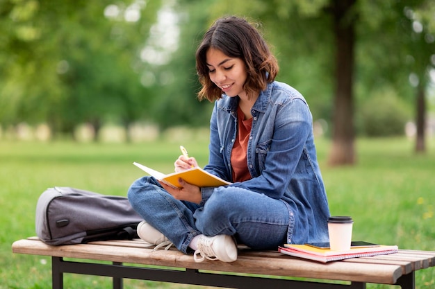 Arabische Studentin schreibt in Notizbuch, während sie auf einer Bank im Park sitzt