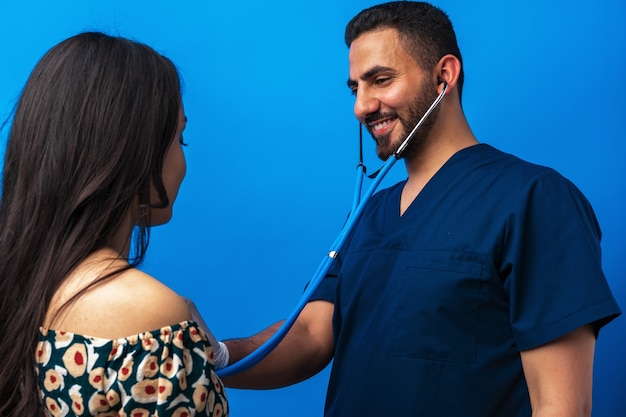 Arabischer Arzt in blauer Uniform, der mit dem Stethoskop steht und dem Herzschlag einer Frau zuhört...