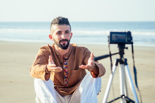 Arabischer Mann, der auf Sand sitzt und Stativvideos am Strand spricht Travel Videographer Lifestyle Content Maker Konzept