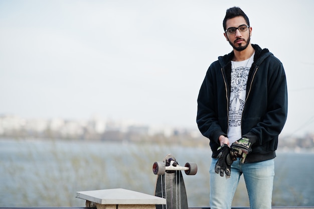 Arabischer Mann im Straßenstil in Brillen mit Longboard, das gegen Fluss gestellt wird.