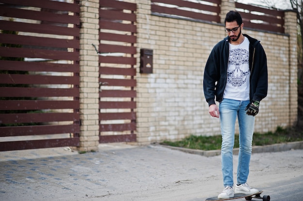 Arabischer Mann im Straßenstil in Brillen mit Longboard Longboarding die Straße hinunter.