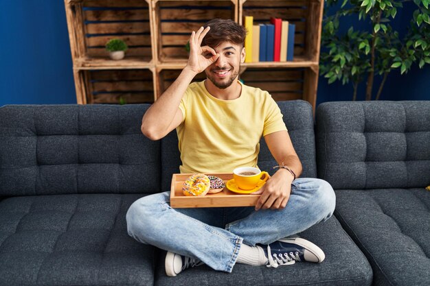 Arabischer Mann mit Bart, der zu Hause auf dem Sofa sitzt und frühstückt, lächelt glücklich und macht ein Ok-Zeichen mit der Hand auf dem Auge, das durch die Finger schaut