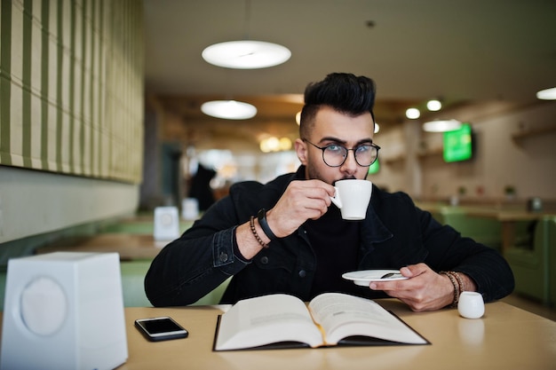 Arabischer Mann trägt schwarze Jeansjacke und Brille, die im Café sitzen, Buch lesen und Kaffee trinken Stilvoller und modischer arabischer Modelltyp