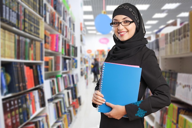 Arabischer Student, der in der Bibliothek steht und Bücher auf seinen Armen hält. Bildungskonzept.