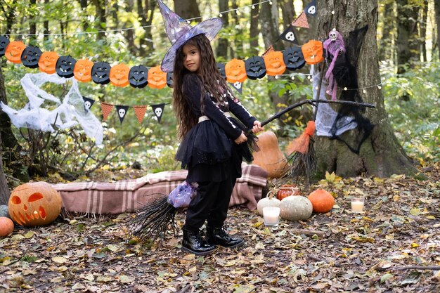 Foto arabisches mädchen in halloween-kostüm und hexenhut mit besen in halloween-dekoration im freien