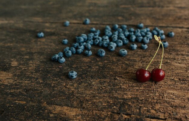 Los arándanos azules frescos están esparcidos sobre una vieja mesa de madera marrón agrietada. Dos cerezas rojas