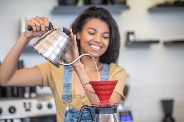 Arbeit, Barista. Konzentrierte junge freudige Frau mit langen dunklen Haaren, die Kaffee mit Tropf in guter Stimmung kocht