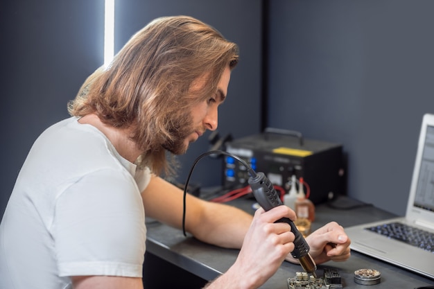 Foto arbeit, mikroschaltung. junger bärtiger konzentrierter mann mit speziellem gerät und mikroschaltung, die am tisch in der werkstatt sitzen