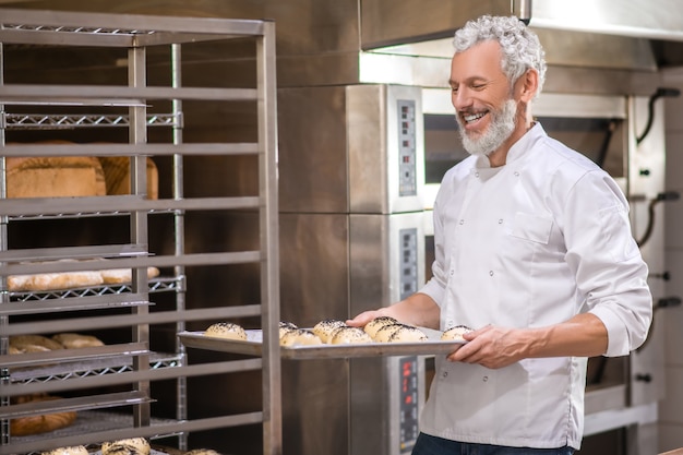 Arbeit, Stimmung. Fröhlicher erwachsener grauhaariger Mann in Uniform mit Tablett der Brötchen, die zum Backen nahe Rack in der Bäckerei vorbereitet werden