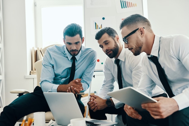 Arbeiten als ein Team. Gruppe junger moderner Männer in formeller Kleidung, die mit Laptop arbeiten, während sie im Büro sitzen