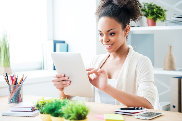 Arbeiten am Tablet. Schöne junge Afrikanerin, die an einem digitalen Tablet arbeitet und lächelt, während sie an ihrem Arbeitsplatz im Büro sitzt