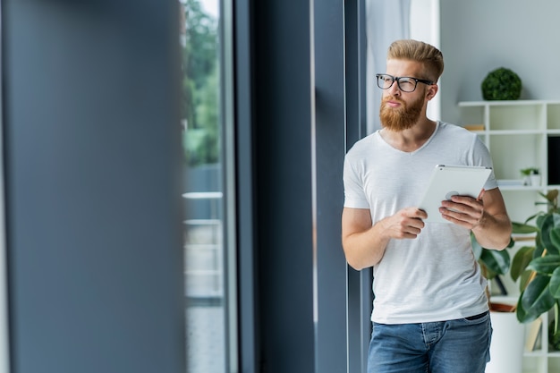 Arbeiten an einem neuen Tablet. Niedrige Winkelansicht des zuversichtlichen jungen Mannes, der auf digitalem Tablett arbeitet, während er vor dem großen Fenster im Büro oder im Café steht.