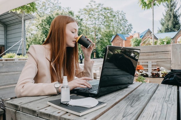 Arbeiten, im Freien, Remote-Arbeit. Redhead Geschäftsfrau, die Kaffee trinkt und draußen am Laptop arbeitet. Junge Geschäftsfrau, die draußen an Laptop arbeitet.