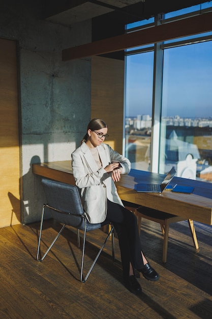 Arbeiten in einem modernen Büro Positive junge Geschäftsfrau in Freizeitkleidung und Brille, die mit einem Mobiltelefon im Büro sitzt, während sie mit einem Laptop an einem Remote-Projekt arbeitet