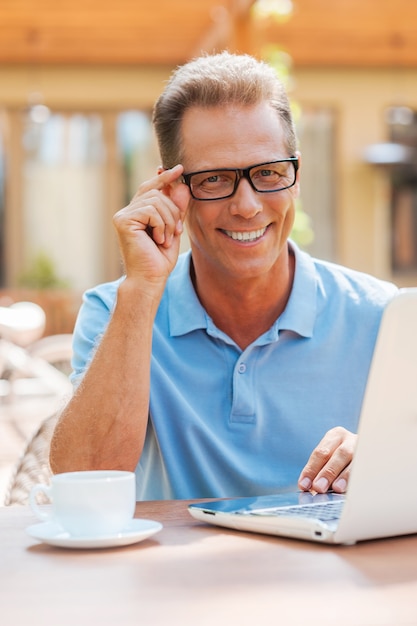 Arbeiten mit Freude. Fröhlicher reifer Mann, der am Laptop arbeitet und lächelt, während er draußen am Tisch mit Haus im Hintergrund sitzt