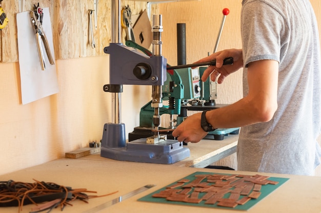 Arbeiten mit Pressmaschine in kleiner DIY Lederwerkstatt. Männliche Hände, die eine Vorrichtung zum Zusammenpressen verwenden, um Nieten in Lederstücke zu stecken