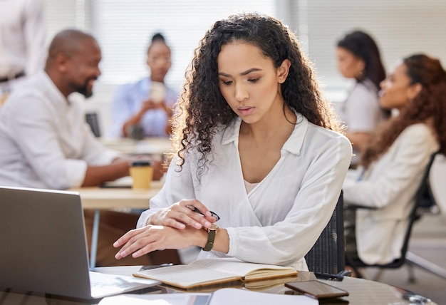 Arbeiten rund um die Uhr, um ihre Fristen einzuhalten Aufnahme einer jungen Geschäftsfrau, die die Zeit überprüft, während sie in einem Büro mit ihren Kollegen im Hintergrund an einem Laptop arbeitet