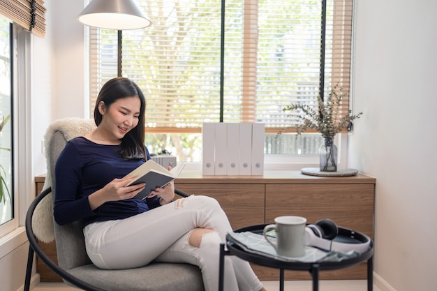 Arbeiten Sie von zu Hause aus. Konzept einer hübschen Frau, die auf einem modernen Stuhl in einem gut eingerichteten, minimalistischen Raum sitzt und ein berühmtes Buch liest.