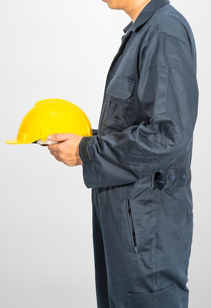 Arbeiter, der im blauen Overall steht und gelben Bauarbeiterhelm auf grauem Hintergrund hält