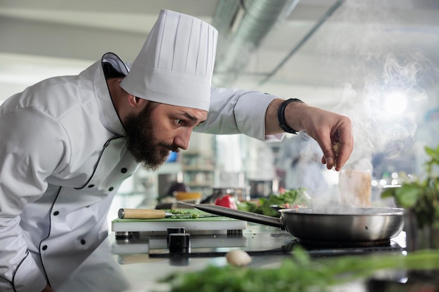 Arbeiter der Lebensmittelindustrie, der Zutaten für das Abendessen in der professionellen Küche zubereitet. Der Chefkoch würzt ein Gourmetgericht in der Pfanne, während er feine Speisen kocht, die beim Abendessen im Restaurant serviert werden.