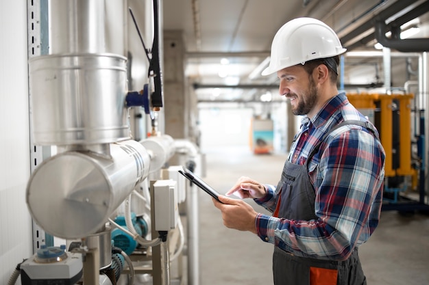 Arbeiter des Wirtschaftsingenieurs, der Tablet-Computer hält und Parameter des Heizungssystems im Fabrikkesselraum einstellt.