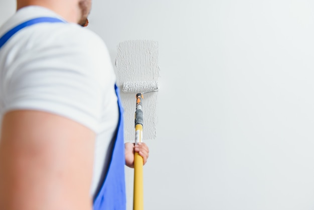 Arbeiter im blauen Overall, der die Wand mit einer Walze streicht, Ansicht von der Seite