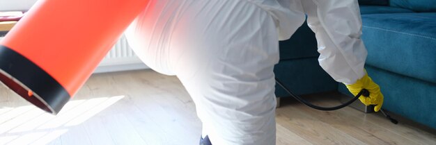 Arbeiter im Schutzanzug behandelt mit Desinfektionslösung auf dem Boden unter dem Sofa in der Wohnung