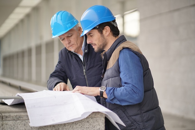 Arbeiter in den Schutzhelmen, die über Plan auf modernem Gebäudeanblick sich beraten