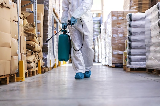 Arbeiter in steriler Uniform mit Gummihandschuhen, die das Sprühgerät mit Desinfektionsmittel halten und um das Lager herum sprühen. Corona-Ausbruchskonzept.