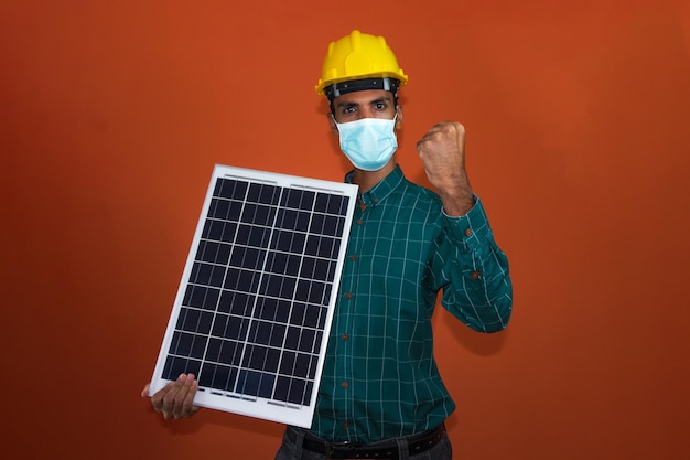 Arbeiter mit Helm und Pandemiemaske, der ein Solar-Photovoltaik-Panel hält, isoliert Solararbeiter isoliert auf orangefarbenem Hintergrund