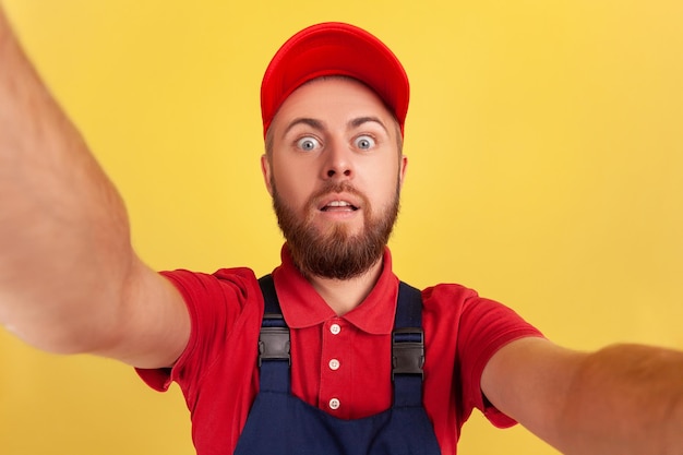 Arbeiter mit roter Mütze und blauer Uniform macht Selfie und blickt mit großen Augen POV in die Kamera