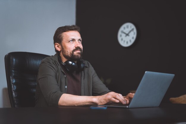 Foto arbeiter träumt, während er im büro sitzt. mann in freizeitkleidung tippt auf dem laptop. allerdings lassen der entfernte blick und die entspannte körperhaltung darauf schließen, dass er in gedanken versunken ist und sich einem moment des träumens hingibt