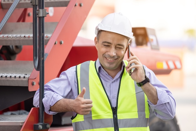 Arbeiterhelm, der auf Mobiltelefon am Versandhof der logistischen Frachtcontainer steht und spricht