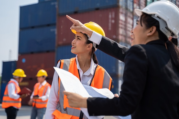 Arbeiterin, die mit Foreman arbeitet und mit einem gelben Helm steht, um das Laden zu kontrollieren und die Qualität von Containern vom Frachtschiff für den Import und Export in der Werft oder im Hafen zu überprüfen