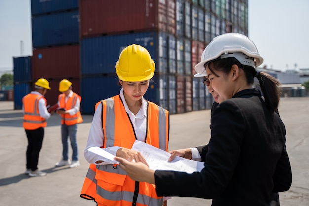 Arbeiterin, die mit Foreman arbeitet und mit einem gelben Helm steht, um das Laden zu kontrollieren und die Qualität von Containern vom Frachtschiff für den Import und Export in der Werft oder im Hafen zu überprüfen