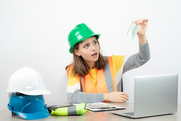 Arbeiterin in orangefarbener Weste und grünem Helm sitzt am Schreibtisch. Foto in hoher Qualität