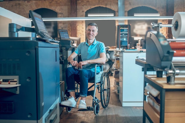 Arbeitsamt. Glücklicher erwachsener Mann in Freizeitkleidung im Rollstuhl mit Blick auf die Kamera in der Nähe von Bürogeräten in beleuchteten Büroräumen