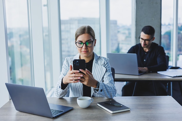 Arbeitsatmosphäre in einem Büro mit großen Fenstern Eine Managerin sitzt an einem Schreibtisch mit Laptop in einem modernen Büro vor dem Hintergrund einer Kollegin