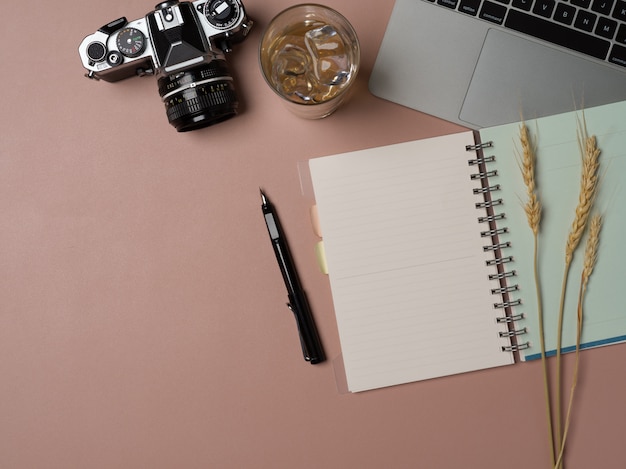 Arbeitsbereich mit geöffnetem Notizbuch, Stift, Laptop, Kamera und Kopierbereich auf rosa Tisch