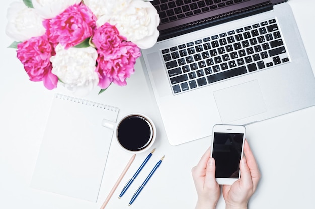 Arbeitsbereich mit Laptop, Handy, Tasse Kaffee und weiblichen Händen, Notizbuch, Stift und rosa und weißen Pfingstrosenblumen auf dem weißen Hintergrund. Flache Lage, Draufsicht. Freiberuflicher Arbeitsplatz